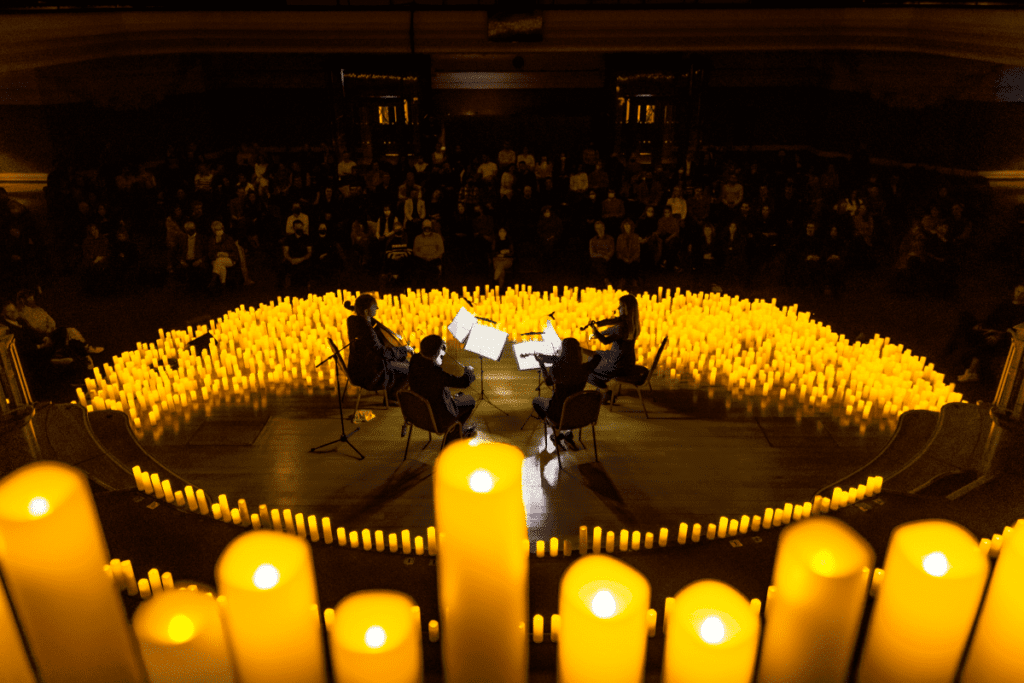 A close up of seven candles with a string quartet playing on a stage covered in candles in the background and the audience visible in the distance.