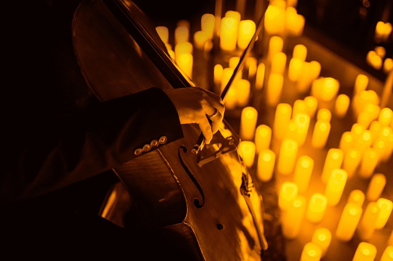 A close up of a hand playing the cello with candles glowing behind it.