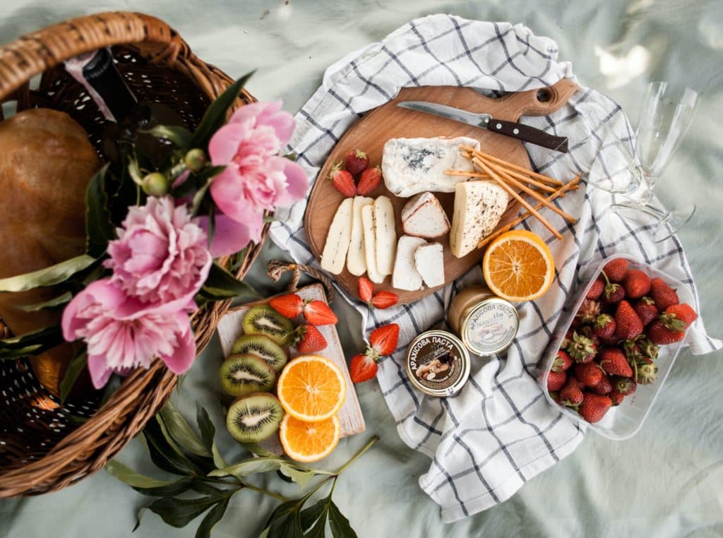 A picnic spread laid out on a blanket with pink peonies, kiwis, oranges, strawberries, and a cheeseboard. 