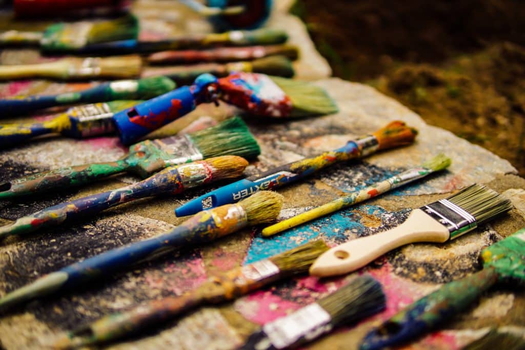 Used paint brushes laid out on a wooden table covered in paint. 