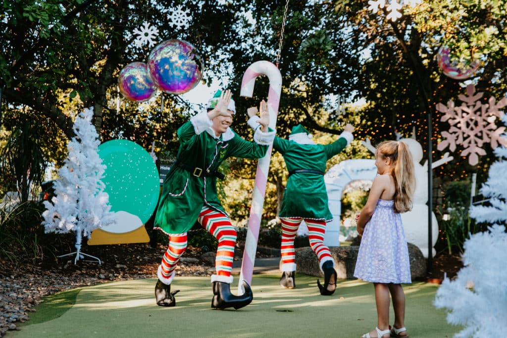 a photo of a girl playing putt putt with two elves at a christmas-themed mini golf course
