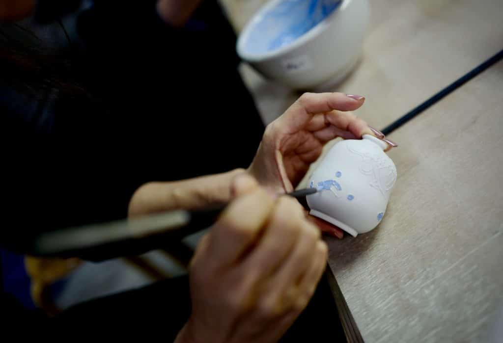 A hand is seen intricately painting a white ceramic piece using blue paint with an already painted clay bowl on the table in the background. 