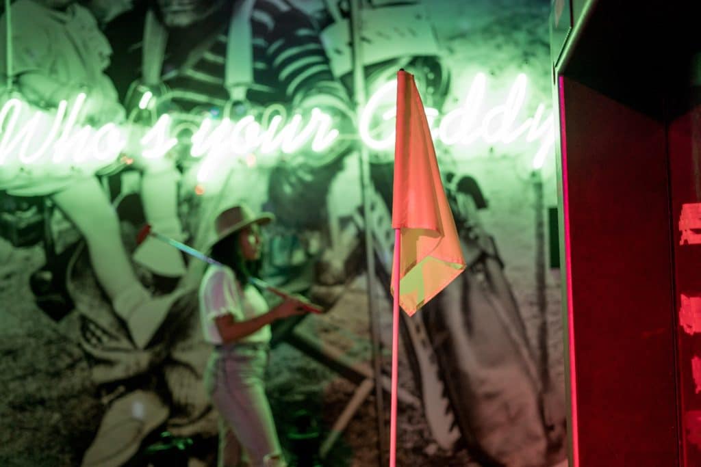 Flag pole in the centre of the image with a woman standing and wearing a hat behind it. There is a mural on the wall in the background with a bright green neon light and it reads, "Who's your daddy".